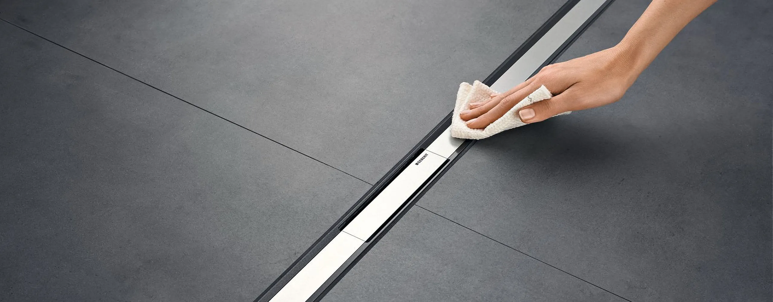 Eine Person wischt eine Duschrinne auf dunklen Fliesen sauber, um die Badezimmerpflege zu demonstrieren. Die glänzende Rinne betont die Sauberkeit.