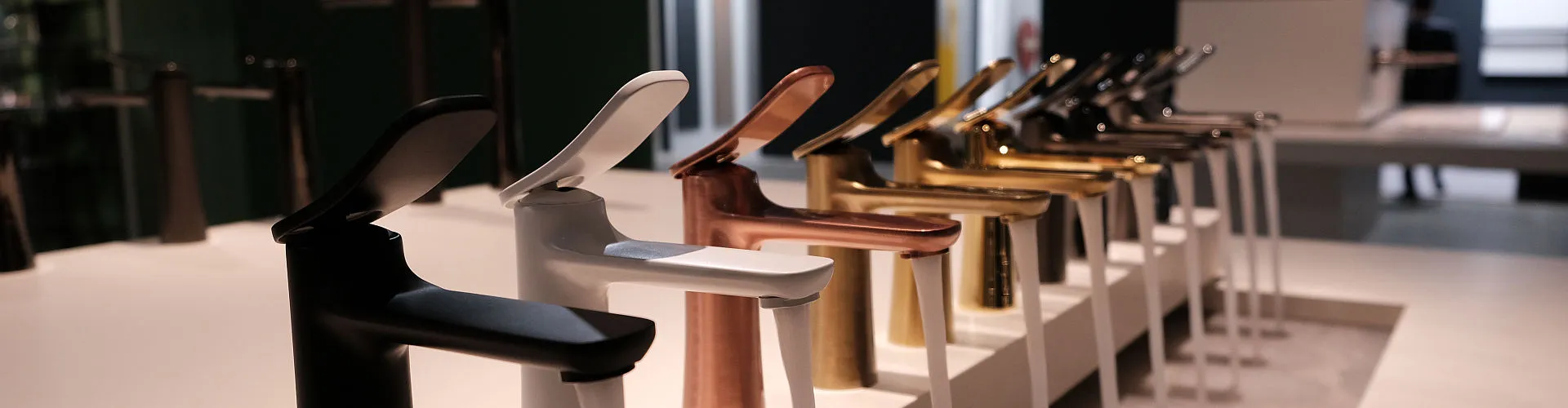 Eine Reihe moderner Wasserhähne der Marke Frattini in verschiedenen Metallic-Finishes, darunter Schwarz, Weiß, Kupfer, Gold und Chrom, ist auf einem stilvollen Waschbeckenpodest ausgestellt. 