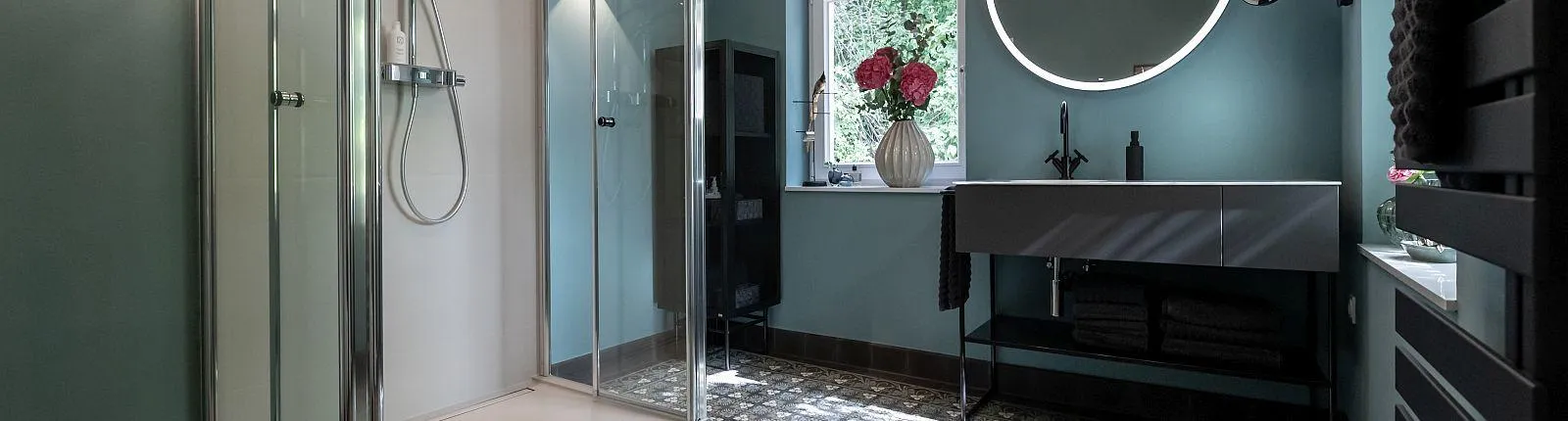 Ein modernes Badezimmer mit Glasdusche, großem beleuchtetem Spiegel und stilvollem Waschbecken vor petrolfarbener Wand.