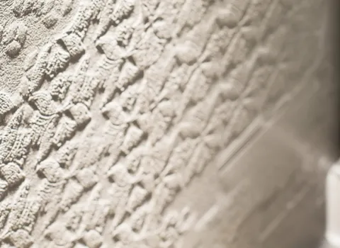 Detailaufnahme einer kunstvoll gemusterten Wandfliese, die verschiedene erhabene Designs und Texturen aufweist. Die helle Farbe der Fliese hebt die komplexen Muster hervor und verleiht dem Raum Tiefe. Im Hintergrund ist ein Teil einer gläsernen Duschabtrennung zu erkennen. Das Bild illustriert die Schönheit und Funktionalität von dekorativen Fliesen.