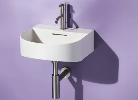 Ein kleines, weißes Wandwaschbecken mit abgerundeten Ecken ist an einer hellvioletten Wand montiert.