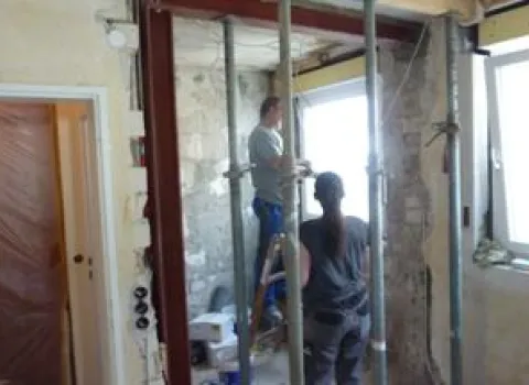 Zwei Handwerker arbeiten an einer offenen Wand in einem im Umbau befindlichen Raum mit freigelegtem Mauerwerk und Stützträgern. 