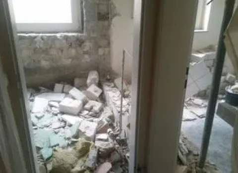 Das Bild zeigt ein Badezimmer im Zustand vollständiger Entkernung, mit herausgerissenen Wand- und Bodenfliesen sowie freigelegtem Mauerwerk. 