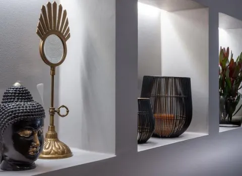 Beleuchtete Wandnischen im Bad mit Buddha-Büste, goldfarbenem Spiegel, schwarzen Windlichtern und Vase mit frischen Blumen.