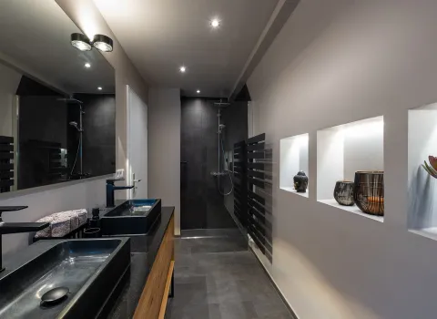 Modernes, schmal geschnittenes Bad mit begehbarer Dusche, schwarzem Doppelwaschtisch, dekorativen Wandnischen und dunklen Fliesen.