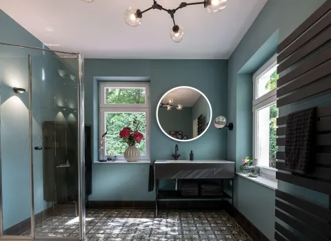 Ein modernes Badezimmer mit stilvollem Design zeigt eine Glasdusche, zwei große Fenster und einen beleuchteten runden Spiegel über einem schwebenden Waschbecken. 