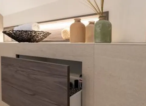 Ein modernes Badezimmer mit integrierter Stauraumlösung zeigt eine geöffnete Schublade in Holzoptik, die Pflegeprodukte enthält.