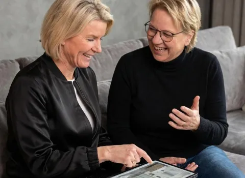 Zwei lächelnde Frauen sitzen auf einem grauen Sofa. Die Frau links, mit blonden Haaren und schwarzer Jacke, zeigt auf ein Tablet, das Inneneinrichtungsoptionen anzeigt. Die Frau rechts, mit blonden Haaren, Brille und schwarzem Rollkragenpullover, lacht und gestikuliert mit ihrer linken Hand.