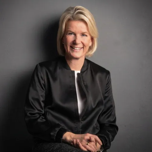 Eine lächelnde Frau mit blonden, kurzen Haaren trägt eine schwarze Satin-Bomberjacke und ein weißes Oberteil. Ihre Hände sind gefaltet und sie sitzt vor einer dunkelgrauen Wand.