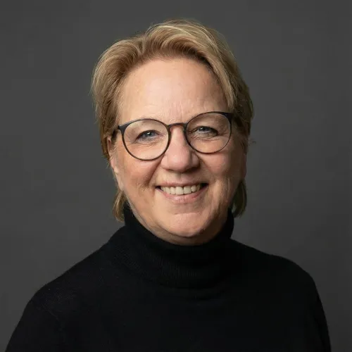 Lächelnde Frau mit kurzen, blonden Haaren, die eine runde Brille mit dunklem Rahmen und einen schwarzen Rollkragenpullover trägt. Sie blickt direkt in die Kamera.