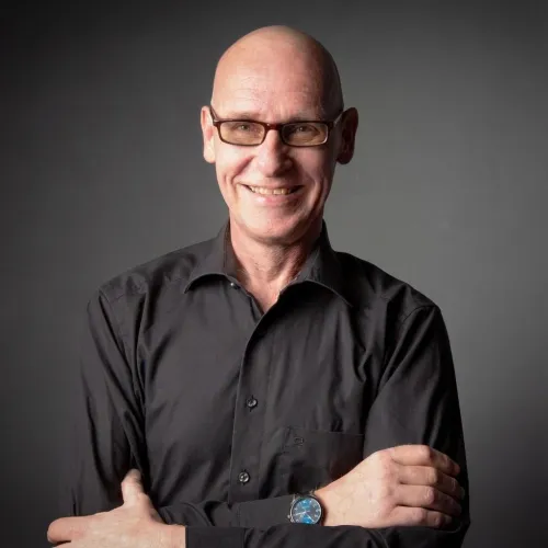 Ein lächelnder, glatzköpfiger Mann mit braunen Brillengestellen und einem schwarzen Hemd, die Arme verschränkt, trägt eine Uhr mit blauem Zifferblatt am linken Handgelenk.