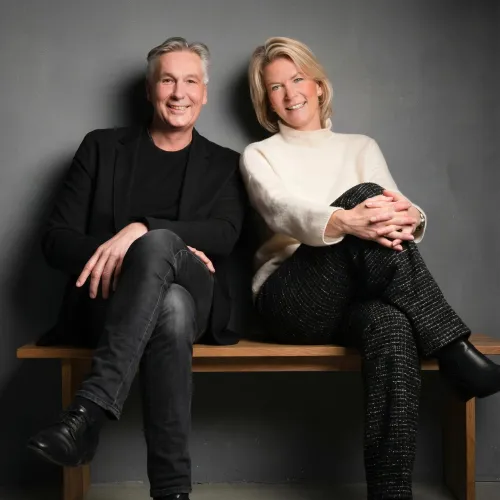 Zwei lächelnde Personen sitzen auf einer Holzbank. Links ein Mann mit grauem Haar, schwarzem Blazer und dunkler Jeans. Rechts eine Frau mit blondem Haar, cremefarbenem Rollkragenpullover und gemusterter Hose. Hintergrund ist eine dunkelgraue Wand.