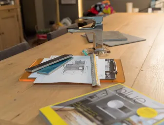Auf einem großen Holztisch liegen ein Badezimmer-Grundriss, Fliesenmuster, ein Maßstab und eine moderne Chromarmatur.