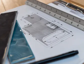 Ein detaillierter Badezimmer-Grundriss liegt auf einem Tisch, umgeben von zwei farbigen Fliesenmustern, einem Lineal und einem schwarzen Stift. 