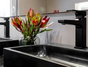 Detailansicht eines modernen schwarzen Waschbeckens mit schwarzer Armatur und Vase mit roten Blüten.