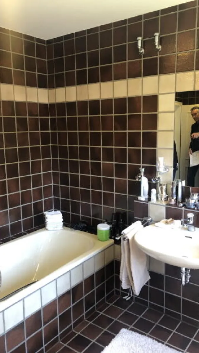 Das Bild zeigt ein Tageslichtbad mit braunen Wandfliesen im 70er-Jahre-Stil, ausgestattet mit zwei Waschbecken und einem Bidet.
