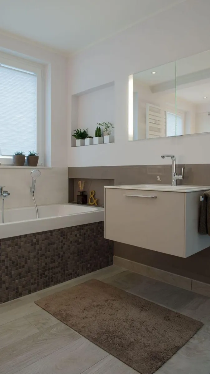 Ein modernes Badezimmer mit Badewanne, die mit dunklen Mosaikfliesen verkleidet ist, daneben ein wandhängender Waschtisch mit eleganter Armatur.