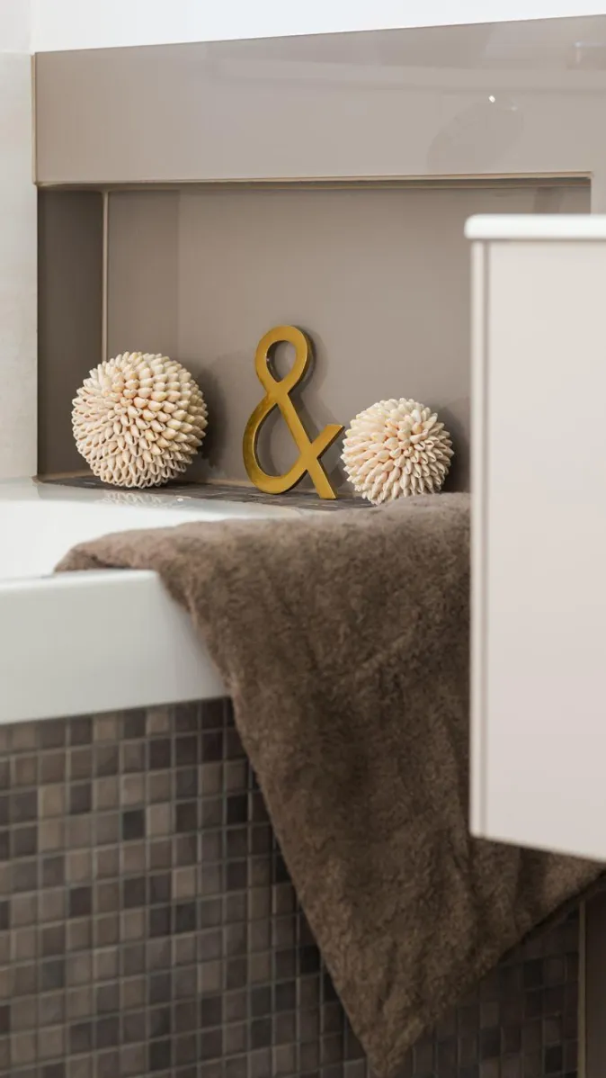 Detailaufnahme einer Badewannenecke mit braunem Handtuch, dunkler Mosaikverkleidung und dekorativer Nische. 