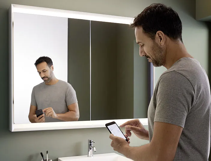 Mann mit Smartphone steht vor einem Badezimmer-Leuchtspiegelschrank der Serie One von Geberit.