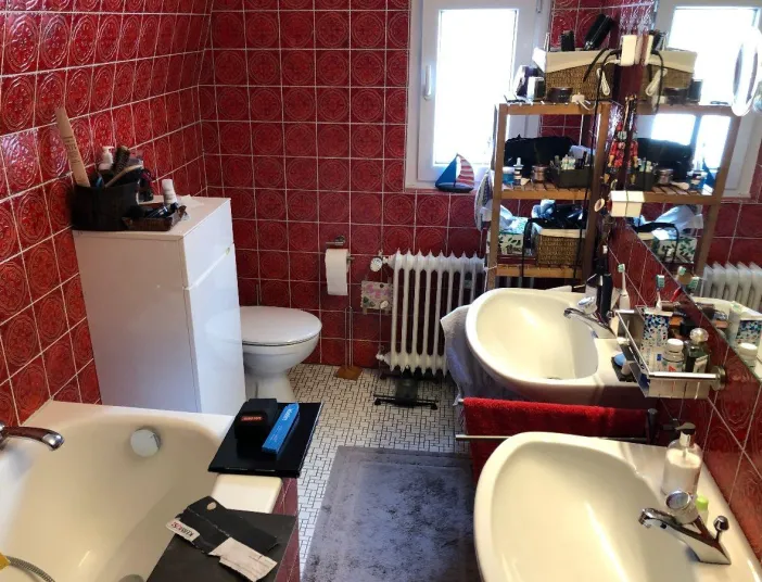 Altes Badezimmer mit roten Retro-Fliesen, Doppelwaschbecken, Badewanne, Heizkörper und offenen Regalen.