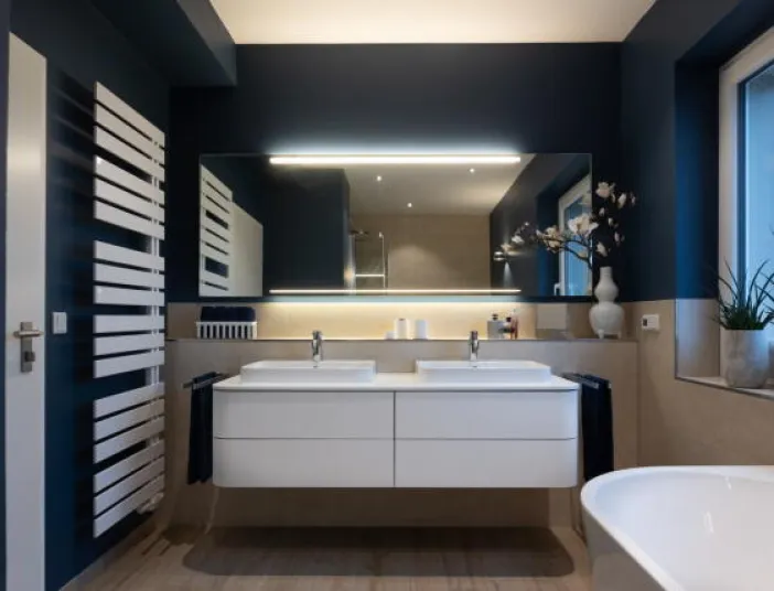 Ein modernes Badezimmer mit dunkelblauen Wänden und klaren Linien präsentiert sich mit einem Doppelwaschbecken unter einem großflächigen Spiegel mit integrierter LED-Beleuchtung. 