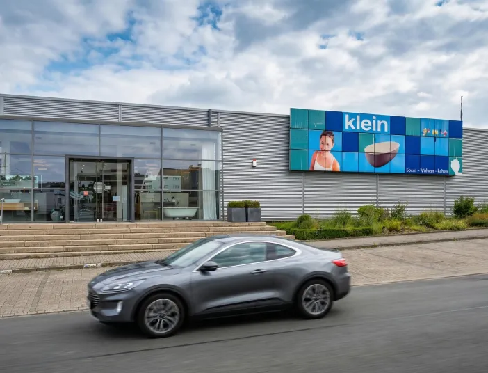 Grauer SUV fährt auf einer Straße vor einem modernen Gebäude. Das Gebäude hat eine Glasfassade, eine Steintreppe zum Eingang und eine horizontale Metallverkleidung. An der Fassade ist eine große Digitalanzeige mit dem Schriftzug "klein", dem Porträt eines Mädchens und einem beigen Waschbecken zu sehen. Im Inneren des Gebäudes sind eine weiße Badewanne und ein Waschtisch sichtbar.